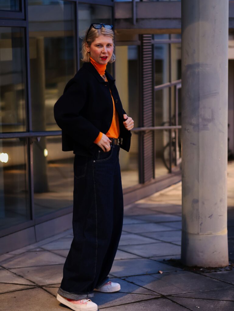 Conny Doll stands on an illuminated city path, combining barrel jeans with a checked quilted jacket, black wool jacket, orange turtleneck, cap and black handbag