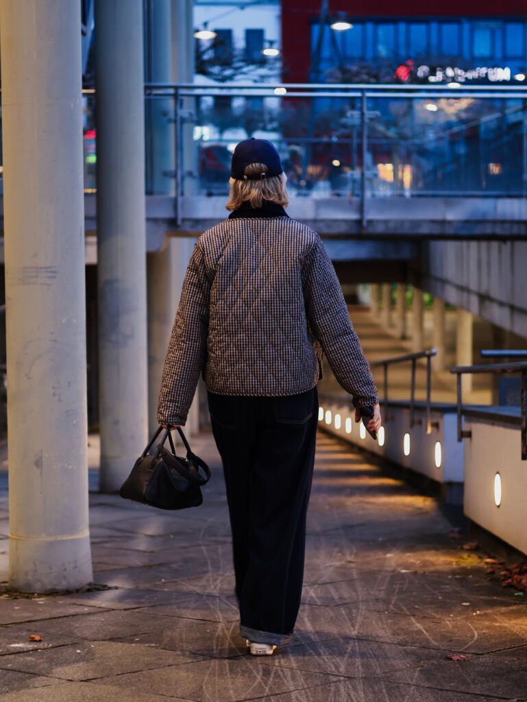 Back view: Conny Doll walks along an illuminated city path, barrel jeans combined with a checked quilted jacket and cap, black handbag
